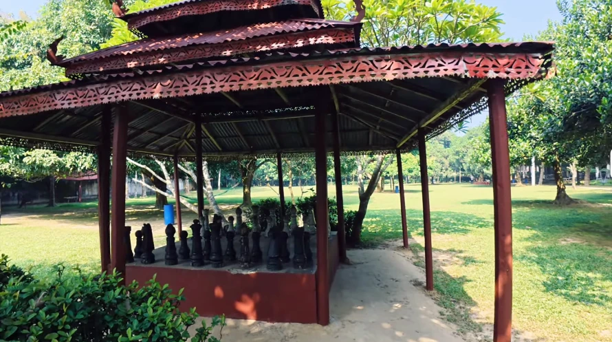 Best Resorts for Couples in Gazipur, Dhaka 26 Wooden Pavilion With Intricate Red Roof Housing Large Chess Pieces At Nuhash Polli In Gazipur, Bangladesh, Surrounded By A Green Open Field And Trees.