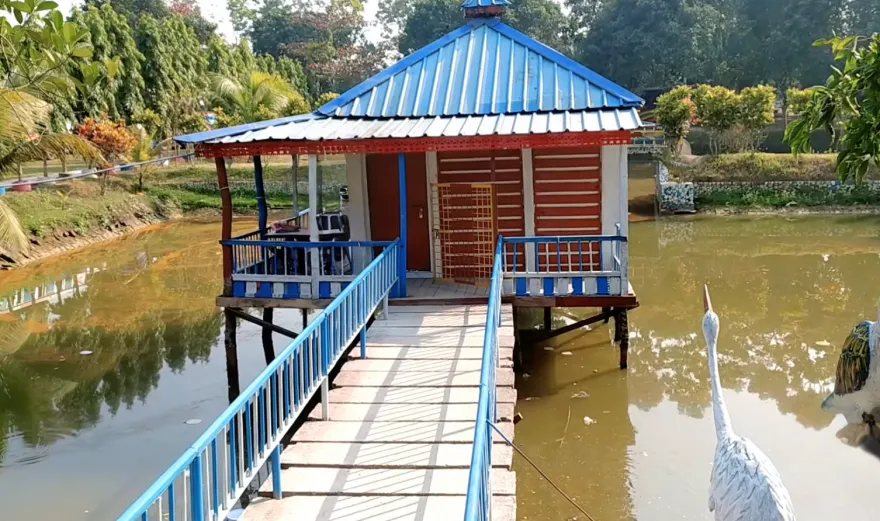 Best Resorts for Couples in Gazipur, Dhaka 18 Beautiful Lakeside Cottage At Sabah Garden Resort In Gazipur, Featuring A Blue Metal Roof, Wooden Walkway Over Water, And Serene Natural Surroundings With Greenery And Decorative Bird Sculptures.