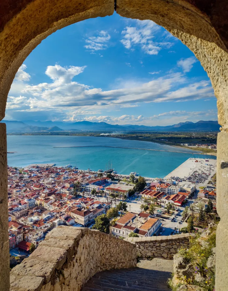 When Is the Best Time of Year to Travel Globally? 3 A Stunning Panoramic View Of A Coastal Town In Europe With Red-Tiled Roofs, A Sparkling Blue Bay, And Mountains In The Distance, Seen Through A Stone Archway.
